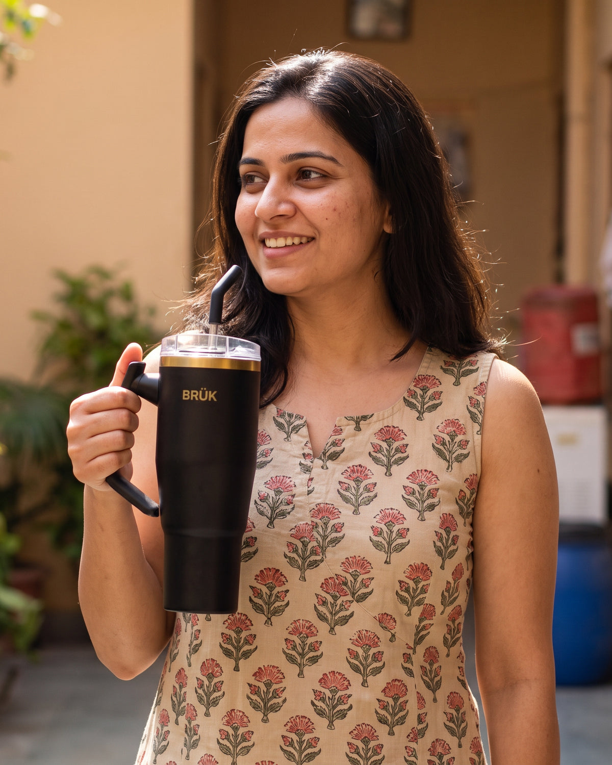 A woman holding a black insulated tumbler with a straw and the brand name BRÜK printed in gold, wearing a beige sleeveless dress with a red and green floral pattern.