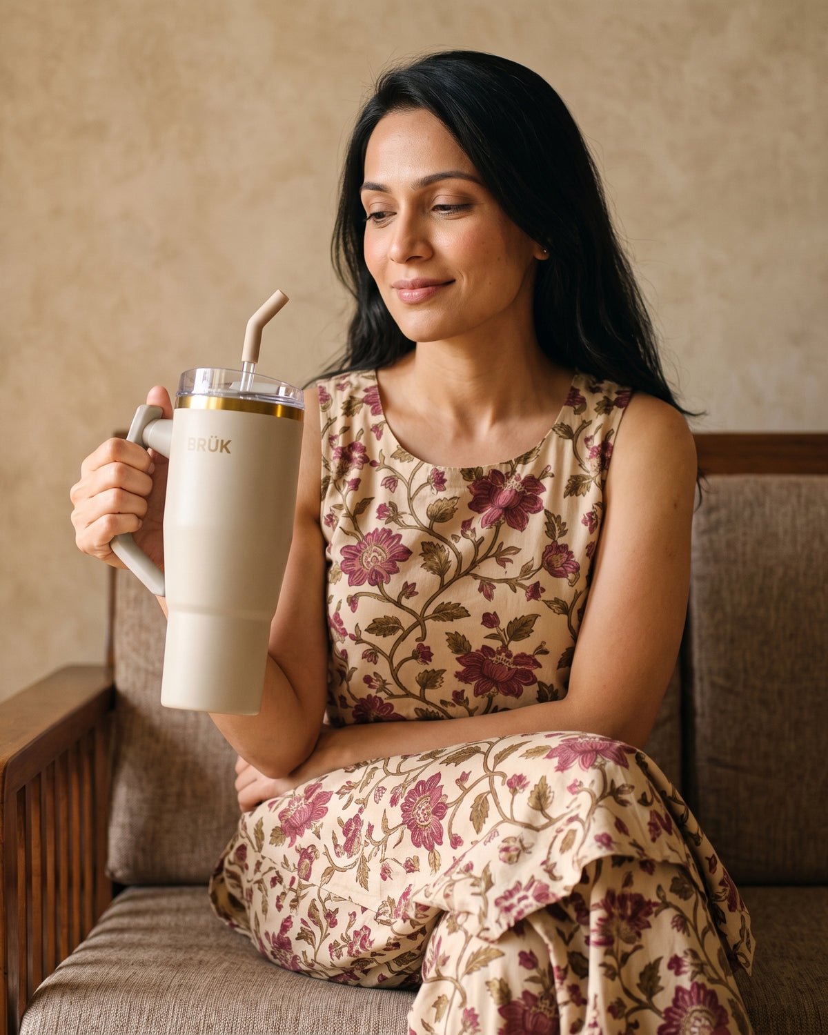 A woman sits on a brown upholstered bench holding a large beige handled tumbler with a clear lid and straw. She wears a sleeveless beige dress with a burgundy floral print.