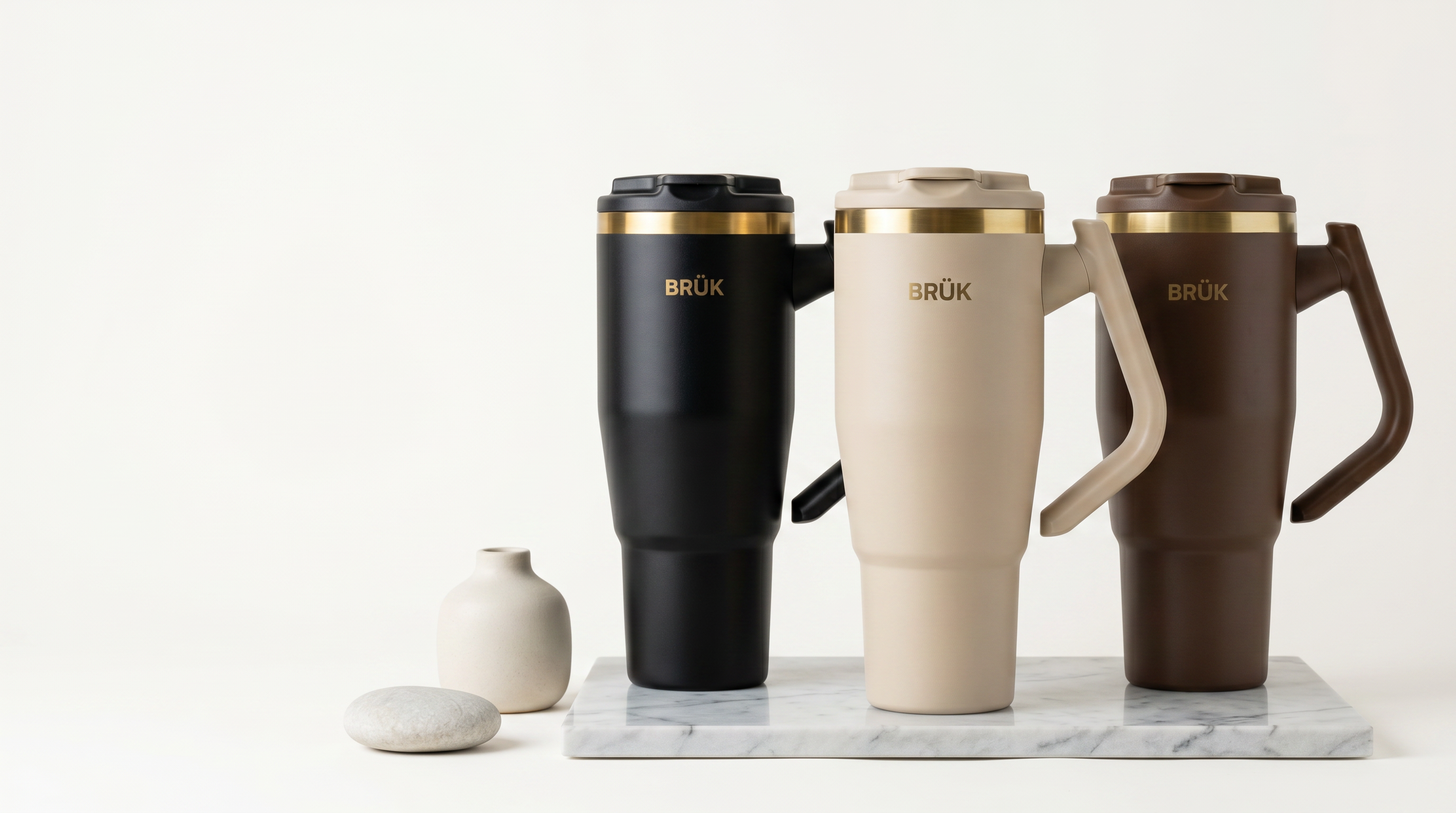 Three BRöK insulated travel mugs in black, beige, and brown on a marble surface with a white background.