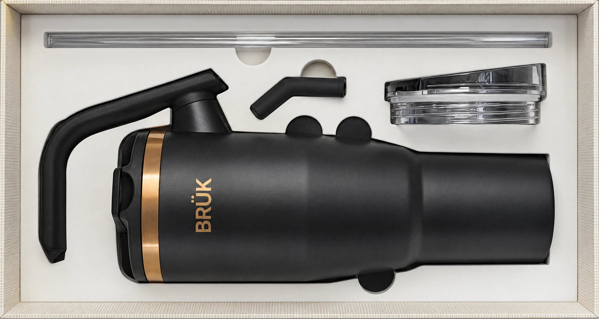 A matte black insulated travel mug with a gold band and gold BRÜK logo, displayed inside its cream-colored presentation box with accessories.