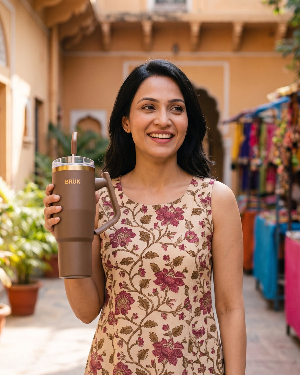 A woman smiling and holding the BRÜK Signature 1.2L Tumbler in Cocoa with a straw lid and gold band.