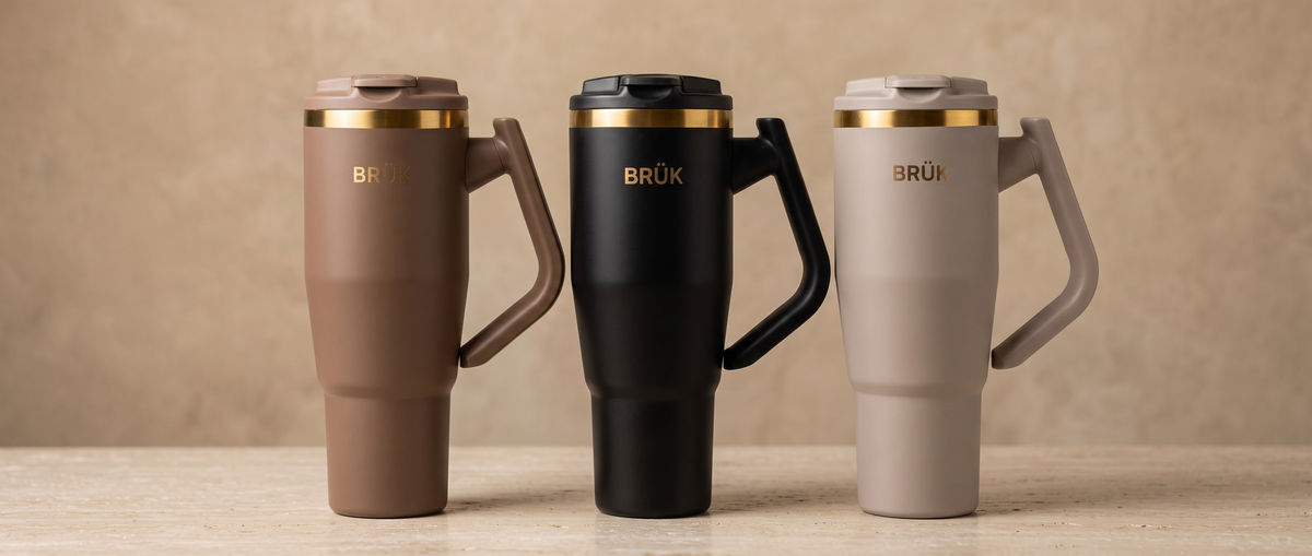 The BRÜK Signature 1.2L Tumbler in Cocoa, Black and Beige side by side, each with its gold band, on a warm stone surface.
